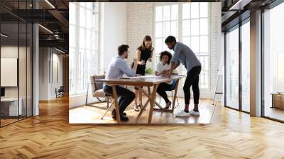 Full length motivated young diverse teammates gathering near table in modern office, working on corporate growth strategy development, analyzing paper marketing research report, discussing ideas. Wall mural