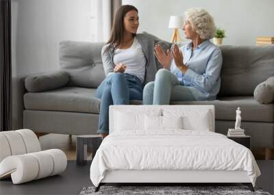 Full length young happy beautiful woman sitting on comfortable couch with smiling middle aged 60s mother, enjoying pleasant conversation in living room, discussing sharing life moments indoors. Wall mural