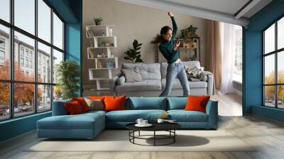 Full of energy. Active young latin lady dance at living room alone do fitness exercise by live music from cellphone. Energetic joyful millennial woman renter tenant jumping having fun at home interior Wall mural