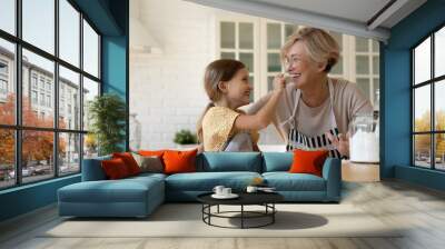 Funny baking. Joyful little girl having fun with retired older grandma when cooking pastries. Laughing elderly granny and cute small grandchild playing at kitchen painting one another noses with flour Wall mural