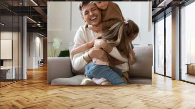 Funny little girls hugging cuddling smiling laughing father sitting on comfortable couch in living room, excited father having fun with two adorable little daughters, enjoying free time together Wall mural