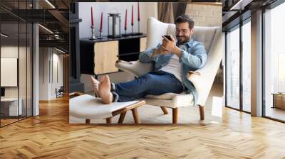 Handsome 30s single man relaxing on comfy armchair put feet on footstool holding in hands smartphone spend weekend alone in cozy cottage house. No stress, home owner, modern tech usage, fun concept Wall mural