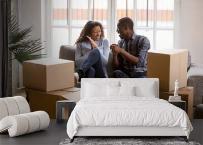 Happy african american couple feeling excited on moving day, black family tenants laughing celebrating relocation sitting on sofa with boxes, home owners having fun in new house together concept Wall mural