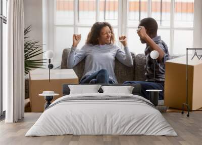 Happy African American couple in love celebrating moving in new house, sitting together on couch with raised hands, cardboard boxes with belongings, start living together, homeowners in new apartment Wall mural