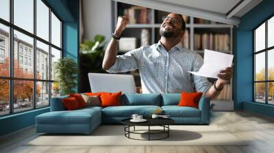 Happy African American man wearing glasses excited by good news, holding letter, sitting at desk, showing yes gesture, celebrating reward, job promotion or lottery win, student received exam results Wall mural