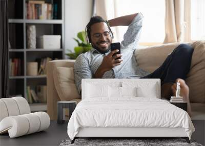 Happy African American man wearing headphones using phone, lying on cozy couch at home, smiling young male enjoying listening to favorite music online, watching video, having fun with smartphone Wall mural