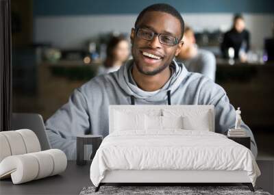 Happy african casual man in glasses sits at cafe table looking at camera, cheerful black student posing studying with book in coffee house, smiling businessman works on laptop in cafeteria, portrait Wall mural