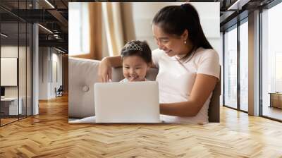 Happy Asian young mother and little daughter using laptop together, looking at screen, sitting on cozy couch at home, smiling mum and cute toddler girl child wearing princess diadem having fun Wall mural