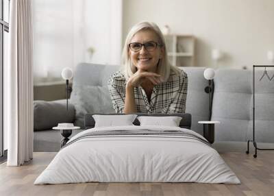 Happy attractive blonde mature woman in stylish eyeglasses sitting on couch, looking at camera, posing with cheerful, positive face, touching chin. Senior pensioner in elegant glasses portrait Wall mural