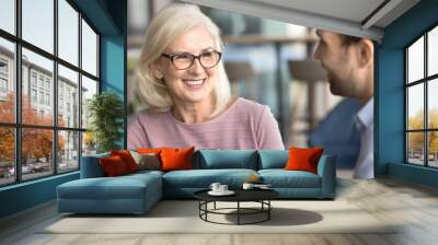 Happy attractive senior businesswoman talking to younger colleague man on business meeting, smiling, laughing, enjoying networking, discussing cooperation, teamwork, partnership Wall mural