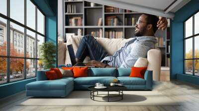 Happy biracial young man relax on comfortable sofa at home working on modern computer gadget, smiling African American millennial male rest on cozy couch in living room using laptop device Wall mural