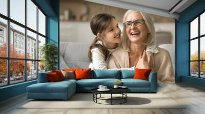 Happy blonde grandma and cheerful granddaughter child playing active games on home sofa. Grandmother and grandkid enjoying funny leisure on family meeting, having fun, hugging on couch Wall mural