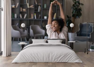 Happy carefree African American woman stretching hands at workplace, leaning back in comfortable chair, student or freelancer relaxing enjoying break after work done, sitting at desk with laptop Wall mural