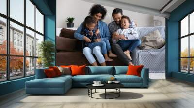 Happy caring young African American parents having fun tickling adorable small children siblings, enjoying entertaining domestic weekend activity, playing together at home, family relations concept. Wall mural