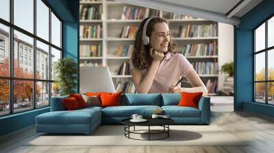 Happy college student girl in wireless headphones enjoying studying in library, sitting at laptop, open books, working on essay, article, writing notes, looking away, thinking, smiling, laughing Wall mural