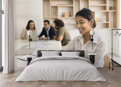 Happy confident business professional girl using smartphone over large office co-working table, sitting in meeting room with diverse colleagues talking in background, typing on cell, smiling Wall mural