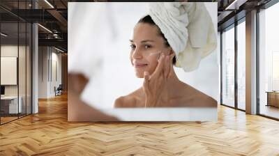 Happy cosmetic product consumer satisfied with moisturizing antiage cream. Young woman spreading nourishing lotion on facial skin after shower, smiling at reflection in mirror. Beauty care concept Wall mural