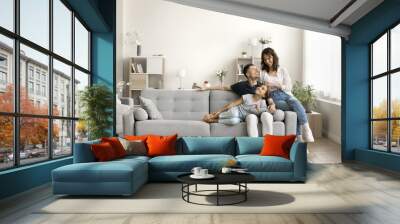 Happy couple of young parents and little daughter kid sitting and relaxing on sofa, resting on comfortable couch in modern cozy home interior, talking, laughing, smiling. Full length wide shot Wall mural