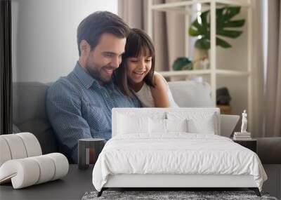 Happy father and little daughter laughing watching funny video using laptop, cheerful dad and child girl having fun playing online game on sofa together, daddy with kid relaxing at home with computer Wall mural