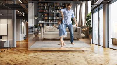 Happy father dancing with adorable little daughter wearing dress, holding hands, moving to favorite music in living room, family having fun, smiling dad and cute girl enjoying leisure time together Wall mural