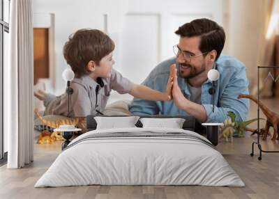 Happy father wearing glasses and little son giving high five, playing with toys, drawing colorful pencils, lying on warm wooden floor with underfloor heating, family having fun at home Wall mural