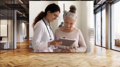 Happy female doctor and senior patient look at tablet screen discuss treatment or therapy on gadget. Smiling woman nurse and mature client use pad device discussing results in modern hospital. Wall mural