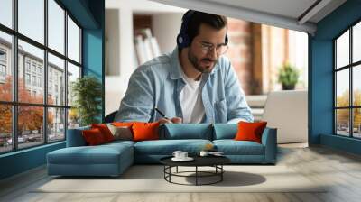 Happy focused young man in eyeglasses wearing wireless headphones, watching educational lecture on computer, studying on online courses distantly, improving professional knowledge, writing notes. Wall mural