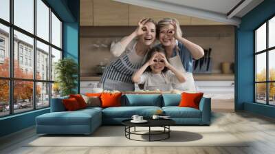 Happy girls and women of different female generations having fun in kitchen, looking at camera through hand finger glasses, smiling, laughing, posing at floury table with dough, bakery ingredients Wall mural