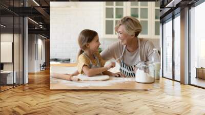 Happy grandmother and adorable little girl cooking homemade pasty cookies in kitchen together, smiling senior woman in glasses with preschool granddaughter having fun, enjoying leisure time Wall mural