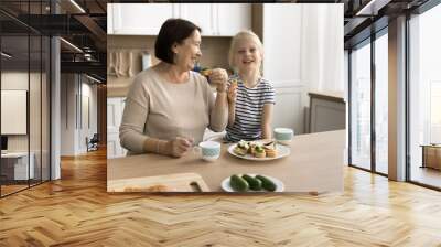 Happy grandmother and cute preschool kid girl having homemade fast food lunch in home kitchen, eating sandwiches, laughing, talking, having fun, looking at camera for casual portrait Wall mural