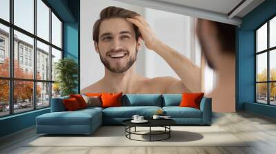 Happy handsome young guy combing smooth brown hair with fingers at mirror in bathroom, looking at reflection with toothy smile. Man satisfied with haircare cosmetic product, enjoying bath routine Wall mural