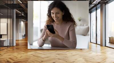 Happy Hispanic young woman sit at desk look at cellphone screen text message online on modern gadget. Smiling Latino millennial female use smartphone talk speak on video call on device at home. Wall mural