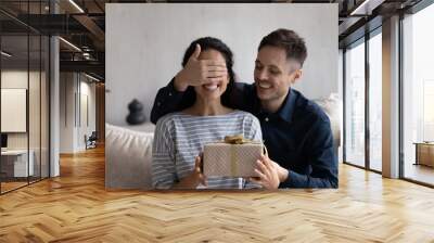 Happy husband giving surprise gift to excited wife. Joyful young man covering girlfriends eyes, giving present box. Millennial couple celebrating birthday, 8 march. Love relationship concept Wall mural