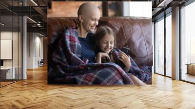 Happy ill mom and little daughter wrapped in plaid relaxing on coach, using mobile phone for playing online game or video call and virtual talk to dad. Mother with cancer spending time with kid Wall mural