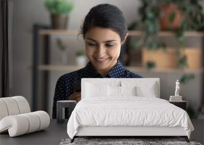 Happy Indian girl smiling at smartphone screen, holding mobile phone. Ethnic millennial woman reading text message on cellphone, typing, chatting online, shopping, browsing on internet, making call Wall mural