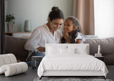 Happy Indian little daughter kid talking to mom, using digital tablet, sitting on sofa at home, smiling, laughing. Mother and child enjoying leisure, playing game on digital gadget Wall mural