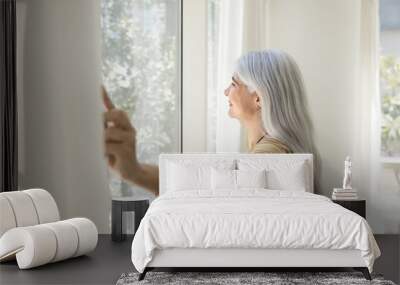 Happy inspired senior Latin woman opening white transparent veils, enjoying morning daylight, looking out of large window, smiling, relaxing in comfortable apartment. Side view, Wall mural