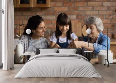 Happy Latin mom and grandma teaching girl to bake in kitchen. Mother, grandmother, kid cooking together, preparing dough for bakery food, mixing egg, flour in bawl on table. Family home activities Wall mural