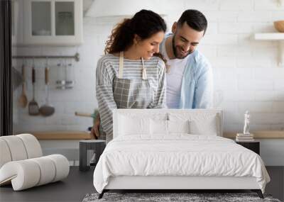 Happy loving couple preparing dinner in modern kitchen together, hugging, standing at table, smiling young woman wearing apron cutting vegetables for salad, spouses enjoying leisure time at home Wall mural