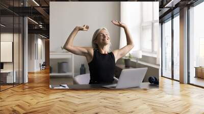 Happy middle aged business woman sitting at desk with eyes closed, rising hands, stretch back after workday finishing, satisfied with good job done, plan weekend, feel successful completion of project Wall mural