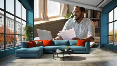 Happy millennial african american businessman in eyewear holding documents, doing paperwork, preparing report or analyzing market research results, working on computer in modern workplace office. Wall mural