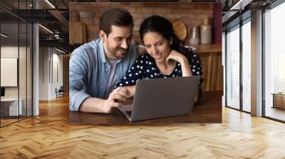 Happy millennial family couple surfing internet on laptop, shopping online together, making order, booking hotel, using app or service. Man and woman pointing at computer screen in kitchen at home Wall mural