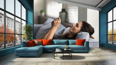 Happy millennial female relax on sofa at home look at cellphone screen texting messaging online. Smiling Caucasian woman rest on couch using smartphone gadget browsing wireless internet or shopping. Wall mural