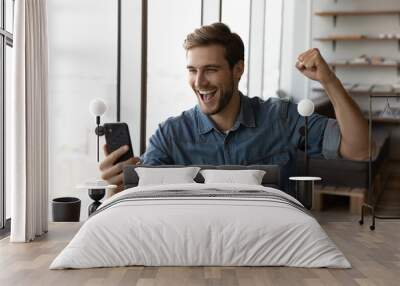 Happy millennial man in casual excited with good news, using smartphone, looking at screen, making winner yes gesture, shouting for joy. Guy reading text message, celebrating win, high result Wall mural