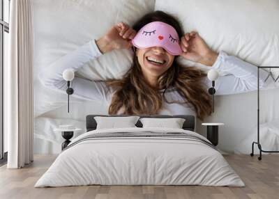 Happy millennial woman lying in bed with sleeping mask. Wall mural