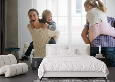 Happy mother laughing piggybacking little son playing with family at home, cheerful parents carrying kids on back having fun together, children boy and girl enjoying active game with mom and dad Wall mural