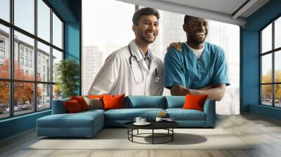 Happy multiethnic young medical colleagues men, cheerful Indian practitioner and African surgeon standing together, looking at camera away, laughing, having fun. Healthcare jobs friends portrait Wall mural