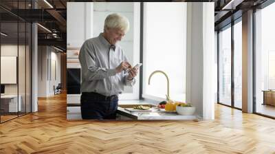 Happy older retired man using mobile phone over kitchen counter, chatting online at table with fresh vegetables for salad, organic food, natural ingredients, browsing recipe on Internet Wall mural