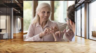 Happy positive old lady with hearing disability speaking sign language to young man, adult son, caregiver, showing hand signs, smiling, laughing, enjoying talk, communication with therapist Wall mural