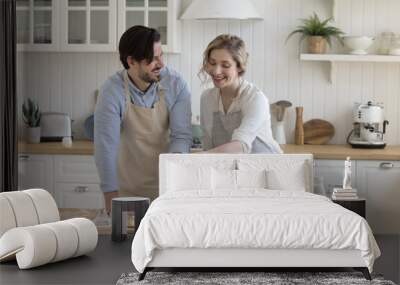 Happy positive young couple in love wearing aprons, enjoying bakery food preparation, baking pies in home kitchen, sifting flour on dough at table with ingredients, talking, smiling Wall mural